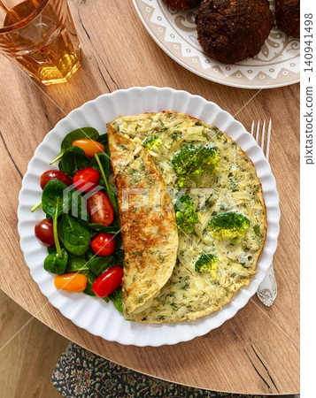 A potato omelette made with eggs and mixed greens is served on a white plate. Spinach, cherry tomatoes, and broccoli are arranged on the side for a healthy breakfast option A potato omelette made with eggs and mixed greens is served on a white plate. Spinach, cherry tomatoes, and broccoli are arranged on the side for a healthy breakfast option 140941498