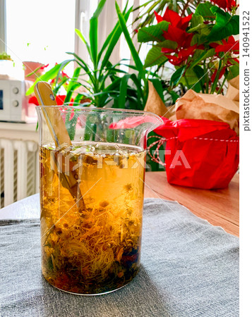A glass container holds a decoction made from chamomile, yarrow, and corn silk. The setup includes a table, plants, and red decorations making the space inviting and bright A glass container holds a decoction made from chamomile, yarrow, and corn silk. The setup includes a table, plants, and red decorations making the space inviting and bright 140941522