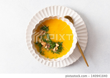 A bowl of pumpkin cream soup sits on a plate. The soup contains nuts and herbs. A yellow checkered napkin lies nearby. This meal can be enjoyed at breakfast or lunch A bowl of pumpkin cream soup sits on a plate. The soup contains nuts and herbs. A yellow checkered napkin lies nearby. This meal can be enjoyed at breakfast or lunch 140941840