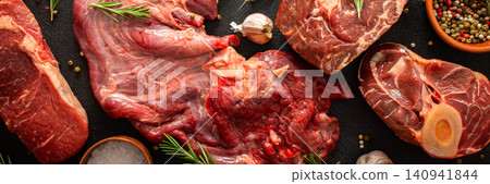 Several types of beef steak are laid out on a dark surface with fresh herbs and spices. Ingredients include garlic, pepper, and oil, showing preparation for cooking in a kitchen, banner Several types of beef steak are laid out on a dark surface with fresh herbs and spices. Ingredients include garlic, pepper, and oil, showing preparation for cooking in a kitchen, banner 140941844