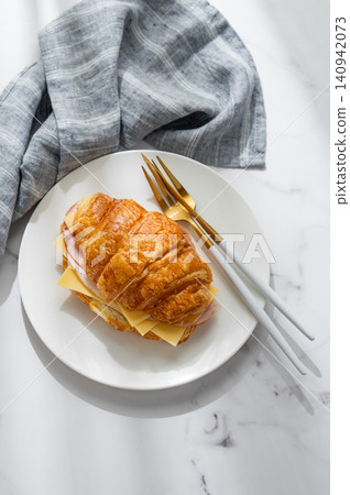 A golden croissant sandwich with ham and cheese on a white plate on a marble background A golden croissant sandwich with ham and cheese on a white plate on a marble background 140942073