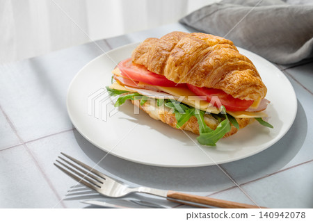 A golden croissant sandwich with ham, cheese, tomato, and arugula on a tile background A golden croissant sandwich with ham, cheese, tomato, and arugula on a tile background 140942078