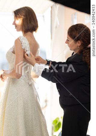 Bride in elegant white wedding dress being assisted by a female stylist adjusting the gown's details in a bright, softly lit dressing room setting Bride in elegant white wedding dress being assisted by a female stylist adjusting the gown's details in a bright, softly lit dressing room setting 140942433