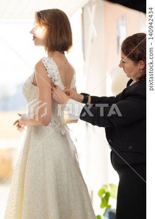 Bride in elegant white wedding dress being assisted by a woman in black attire while preparing for a ceremony in a bright, softly lit indoor setting Bride in elegant white wedding dress being assisted by a woman in black attire while preparing for a ceremony in a bright, softly lit indoor setting 140942434