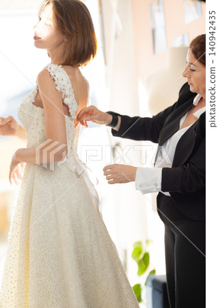 Bride fitting a wedding dress with assistance from a female stylist in a well-lit room, showcasing intricate lace details and soft fabric textures Bride fitting a wedding dress with assistance from a female stylist in a well-lit room, showcasing intricate lace details and soft fabric textures 140942435