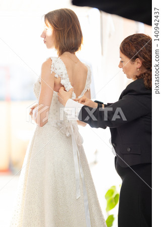 Female model in white wedding dress being assisted by stylist with dress adjustments in a bright, elegant fitting room setting with soft lighting Female model in white wedding dress being assisted by stylist with dress adjustments in a bright, elegant fitting room setting with soft lighting 140942437
