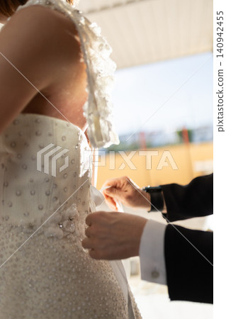 Bride in elegant wedding dress with intricate beadwork adjusts corset while another person assists with the bow in a bright, softly lit indoor setting Bride in elegant wedding dress with intricate beadwork adjusts corset while another person assists with the bow in a bright, softly lit indoor setting 140942455