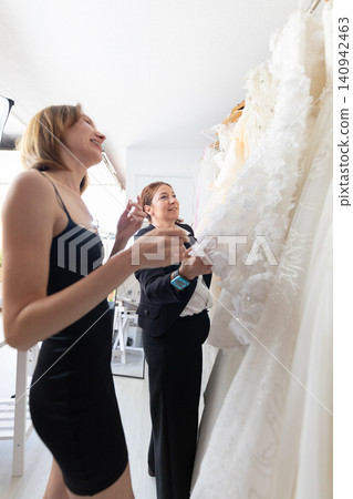 Two women selecting wedding dresses from a rack in a bright boutique, one in a black dress and the other in formal attire, showcasing various white gowns Two women selecting wedding dresses from a rack in a bright boutique, one in a black dress and the other in formal attire, showcasing various white gowns 140942463