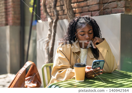 Thoughtful african american female on coffee break in cafe, sitting at table scrolling on smartphone Thoughtful african american female on coffee break in cafe, sitting at table scrolling on smartphone 140942564
