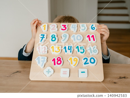 the boy learns to count, holds a board with numbers-puzzles arranged in order from 1 to 20, the boy is 4-5 years old European appearance, at home sits at the table, preparation for school.  140942583