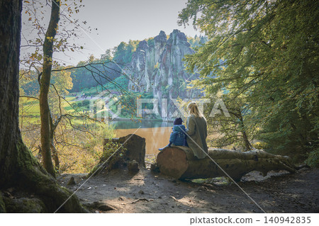Mom and baby sit on a fallen tree trunk with their backs to the camera and look out over the lake Mom and baby sit on a fallen tree trunk with their backs to the camera and look out over the lake 140942835