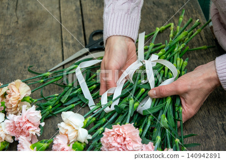 A person arranges pink flowers on a wooden table while tying them with ribbons in a bright workspace A person arranges pink flowers on a wooden table while tying them with ribbons in a bright workspace 140942891