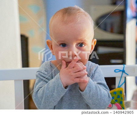 A frightened boy under one year old standing in his crib and holding his hands to his mouth, a boy of European appearance with blue eyes dressed in a gray bodysuit. High quality photo 140942905