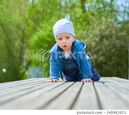 boy up to one year old boy European appearance blue eyes sits in summer on a wooden bench dressed in denim overalls and a light cap . High quality photo 140942913