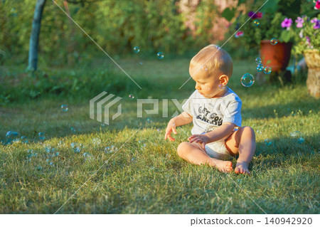 boy one year old boy crawling on the grass in the summer for soap bubbles dressed in a T-shirt and a diaper, the baby is of European appearance blond with blue eyes. High quality photo boy one year old boy crawling on the grass in the summer for soap bubbles dressed in a T-shirt and a diaper, the baby is of European appearance blond with blue eyes. High quality photo 140942920