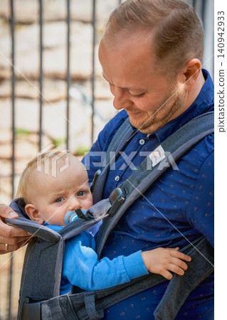 a boy under one year old sits at his father's side in a kangaroo backpack and looks into the camera, European-looking blondes with blue eyes, a baby with a pacifier in his mouth. High quality photo a boy under one year old sits at his father's side in a kangaroo backpack and looks into the camera, European-looking blondes with blue eyes, a baby with a pacifier in his mouth. High quality photo 140942933