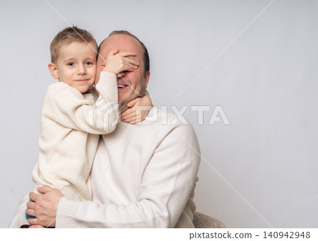 boy 4-5 years old boy sitting on daddy's lap boy covers daddy's eyes with his hand, the boy laughs, both of European appearance blond with blue eyes, studio shooting, dressed in light-colored clothes boy 4-5 years old boy sitting on daddy's lap boy covers daddy's eyes with his hand, the boy laughs, both of European appearance blond with blue eyes, studio shooting, dressed in light-colored clothes 140942948