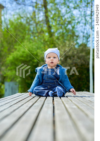 boy up to one year old boy European appearance blue eyes sits in summer on a wooden bench dressed in denim overalls and a light cap . High quality photo boy up to one year old boy European appearance blue eyes sits in summer on a wooden bench dressed in denim overalls and a light cap . High quality photo 140942949