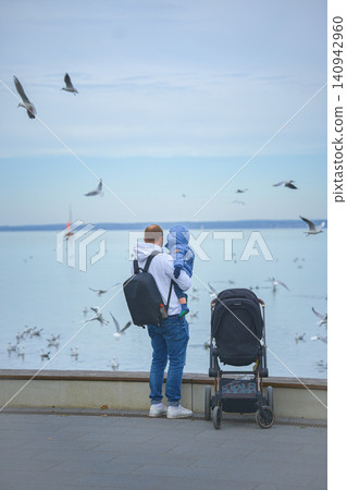 A father holds his one-year-old son in his arms and a stroller stands next to him. They stand with their backs to the camera and look at the lake over which seagulls fly and a small sailboat sails on A father holds his one-year-old son in his arms and a stroller stands next to him. They stand with their backs to the camera and look at the lake over which seagulls fly and a small sailboat sails on 140942960