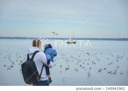 A father holds his one-year-old son in his arms, they stand with their backs to the camera and look at the lake over which seagulls fly and a small sailboat sails on an autumn day. High quality photo 140942962