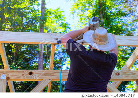 Handy worker works with power tool on wooden beams in backyard surrounded by trees Handy worker works with power tool on wooden beams in backyard surrounded by trees 140943717