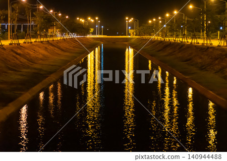 Night canal illuminated by glowing streetlights golden reflections calm water creating an urban landscape featuring symmetry tranquility modern infrastructure Night canal illuminated by glowing streetlights golden reflections calm water creating an urban landscape featuring symmetry tranquility modern infrastructure 140944488