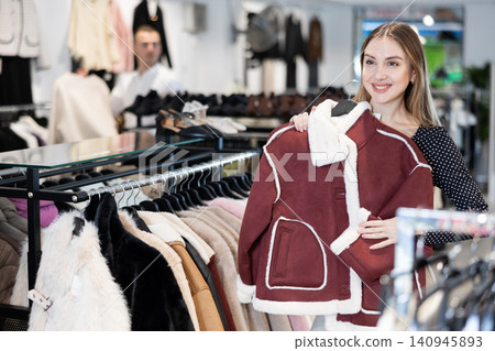 Girl stopped near showcase with outerwear, holds fur coat hanger in hands and examines product Girl stopped near showcase with outerwear, holds fur coat hanger in hands and examines product 140945893