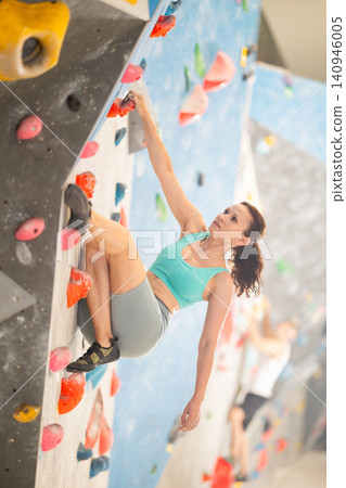 Adult woman is mastering climbing on training wall in gym, side view. Young woman holds on tightly to ledges and strives for top of bouldering route 140946005
