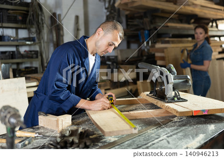 Man employee of carpentry workshop makes measurements of wooden board, counts off set parameters Man employee of carpentry workshop makes measurements of wooden board, counts off set parameters 140946213