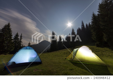 Bright big moon in dark blue cloudy sky over two tourist tents on green grassy forest clearing among tall pine trees on distant mountain background. Tourism, night camping in summer mountains. 140946412