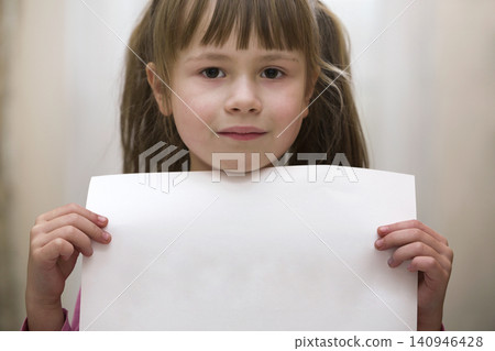 Happy smiling cute child girl holding white copy space sheet of drawing paper. Art education, creativity, advertisement concept. Happy smiling cute child girl holding white copy space sheet of drawing paper. Art education, creativity, advertisement concept. 140946428