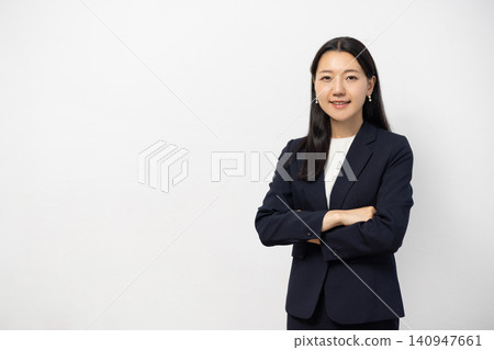 A young Japanese businesswoman stands in front of a white wall, arms crossed, smiling and looking directly at the camera. A young Japanese businesswoman stands in front of a white wall, arms crossed, smiling and looking directly at the camera. 140947661