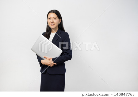 A young Japanese businesswoman smiles at the camera, holding a laptop, in front of a white wall. A young Japanese businesswoman smiles at the camera, holding a laptop, in front of a white wall. 140947763