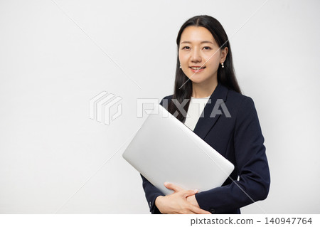 A young Japanese businesswoman smiles at the camera, holding a laptop, in front of a white wall. A young Japanese businesswoman smiles at the camera, holding a laptop, in front of a white wall. 140947764