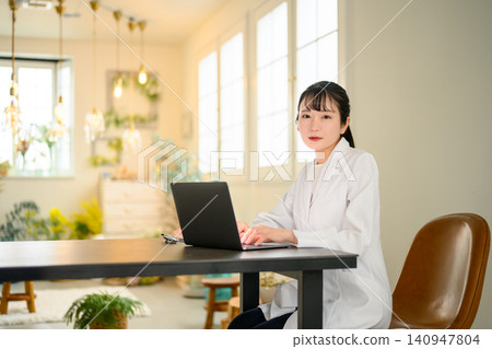 A woman wearing a white coat is operating a laptop computer. 140947804