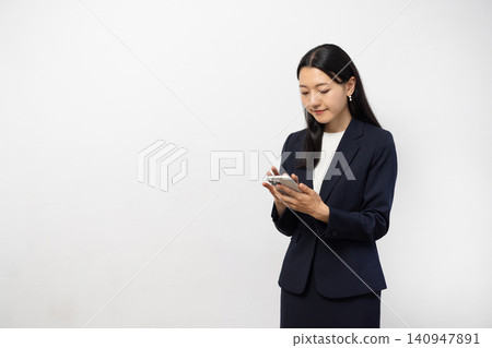 A young Japanese businesswoman is using her smartphone in front of a white wall. A young Japanese businesswoman is using her smartphone in front of a white wall. 140947891