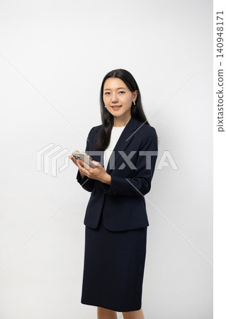 一位年輕的日本女商人手持智慧型手機，站在白牆前，對著鏡頭微笑。 140948171