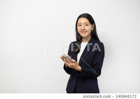 A young Japanese businesswoman smiles at the camera, holding her smartphone, in front of a white wall. A young Japanese businesswoman smiles at the camera, holding her smartphone, in front of a white wall. 140948172