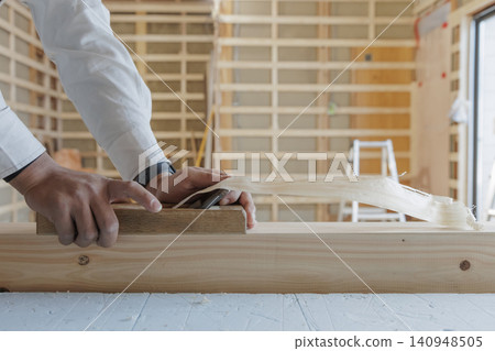 Against the backdrop of a wooden building's framework, a craftsman planes lumber, his hands shaving wood as he finishes the job. Against the backdrop of a wooden building's framework, a craftsman planes lumber, his hands shaving wood as he finishes the job. 140948505