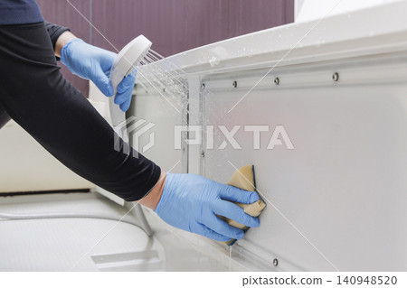 Cleaning staff wearing blue gloves rinse the inside of the bathtub apron with a shower and sponge. Cleaning staff wearing blue gloves rinse the inside of the bathtub apron with a shower and sponge. 140948520