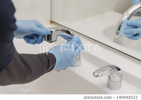 Cleaning staff in blue gloves scrubbing the sink faucet with a sponge; bathroom cleaning reflected in the mirror. Cleaning staff in blue gloves scrubbing the sink faucet with a sponge; bathroom cleaning reflected in the mirror. 140948522