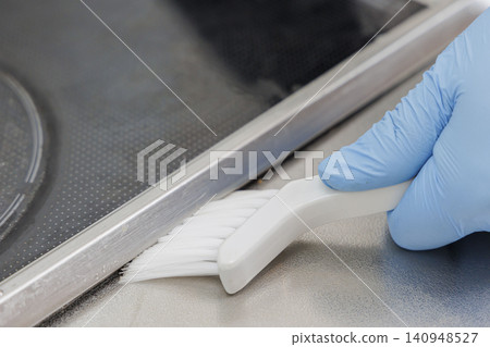 A close-up of someone scrubbing the grooves around the edge of an induction cooktop with a white brush. A close-up of someone scrubbing the grooves around the edge of an induction cooktop with a white brush. 140948527