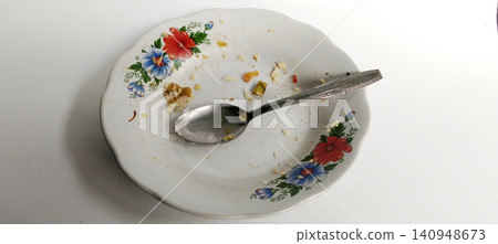 An empty floral-patterned ceramic plate, accompanied by a spoon and food crumbs, set against a white background. An empty floral-patterned ceramic plate, accompanied by a spoon and food crumbs, set against a white background. 140948673