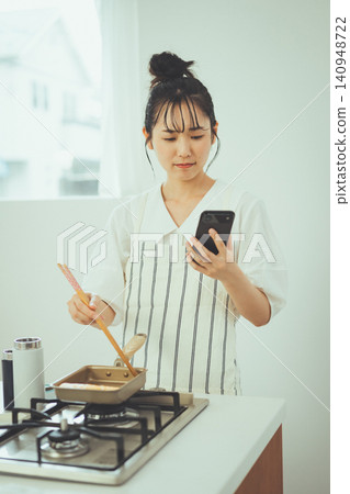 A woman cooking an omelet while looking at her smartphone. 140948722