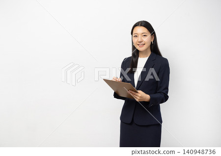 一位年輕的日本女商人對著鏡頭微笑，同時在白牆前的活頁夾上做筆記。 140948736
