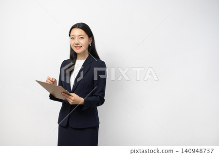 一位年輕的日本女商人對著鏡頭微笑，同時在白牆前的活頁夾上做筆記。 140948737