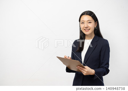 一位年輕的日本女商人對著鏡頭微笑，同時在白牆前的活頁夾上做筆記。 140948738