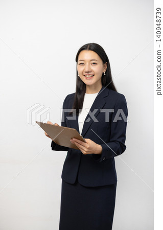 一位年輕的日本女商人對著鏡頭微笑，同時在白牆前的活頁夾上做筆記。 140948739