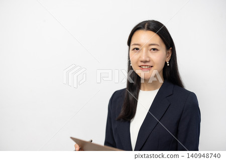 一位年輕的日本女商人對著鏡頭微笑，同時在白牆前的活頁夾上做筆記。 140948740