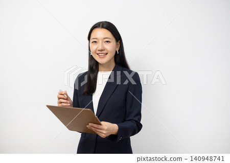 一位年輕的日本女商人對著鏡頭微笑，同時在白牆前的活頁夾上做筆記。 140948741